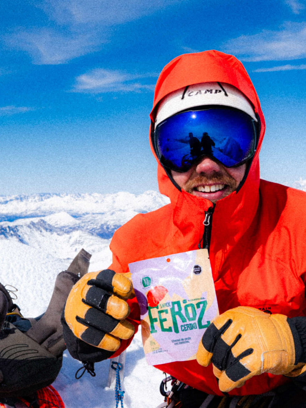 Un montañista con una chaqueta naranja brillante y gafas reflectantes azules sonríe mientras sostiene un paquete de "Feroz", el mejor charqui artesanal de cerdo y vacuno, elaborado por La Fiambrería. De fondo, se aprecian cumbres nevadas y un cielo azul despejado. A su lado, se ven una mochila y equipo de escalada.